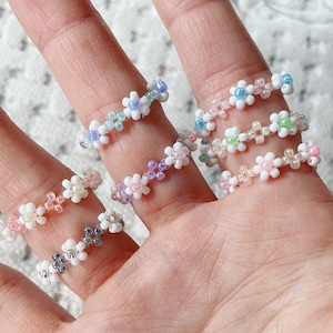 May include: Five colorful beaded rings with flower designs. The rings are made of small beads in shades of pink, blue, white, and purple.