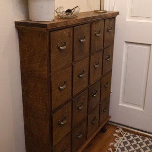 May include: A wooden cabinet with numerous drawers, each with brass handles. The cabinet is stained a warm brown and stands on a wooden floor. A decorative candle holder and a vase with dried flowers are on top.