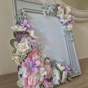 May include: A light blue picture frame adorned with a floral arrangement of pink, white, and purple flowers. The flowers are arranged around the frame's edges, creating a delicate and romantic look.