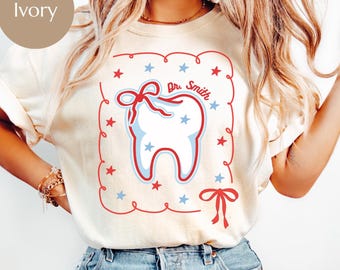 May include: Ivory-colored t-shirt featuring a cartoon tooth design with a red bow and stars. The design includes the text "Dr. Smith" within a decorative frame. The shirt is a casual style.