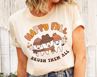 May include: A beige t-shirt with a graphic of three cartoon teeth wearing fall-themed hats and scarves. The text "Happy Fall Brush Them All" is printed below the teeth.