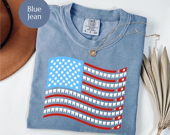 May include: Blue jean colored t-shirt with a graphic of the American flag, where the stripes are replaced with rows of teeth. The upper left corner features a blue square with white stars. The shirt has a crew neck and short sleeves.