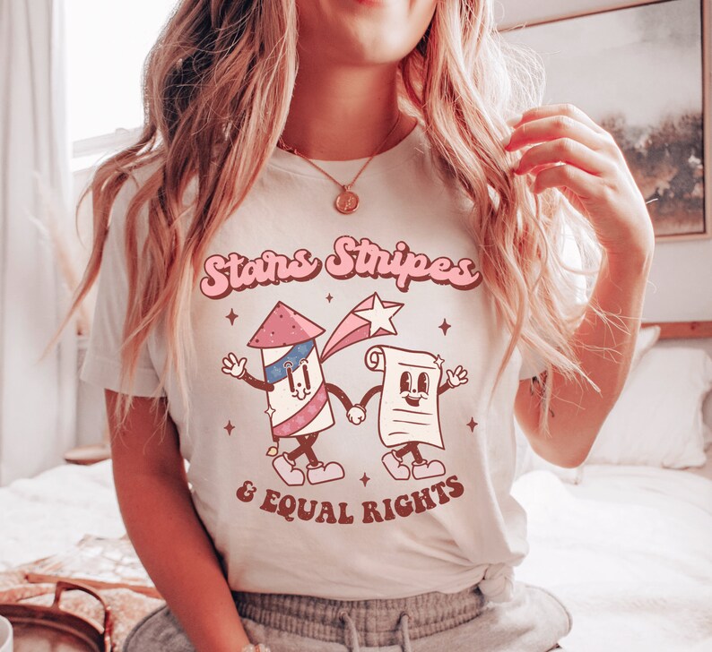 May include: A light-colored t-shirt featuring a retro design with the words "Stars Stripes & Equal Rights." The graphic includes cartoon illustrations of a firework and a scroll, both with smiling faces. The design uses red, white, and blue colors.