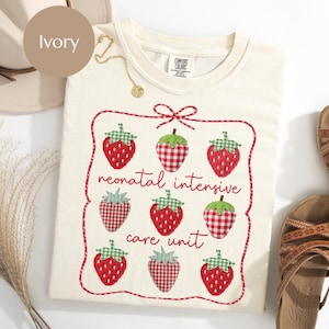 May include: Ivory-colored sweatshirt with a strawberry design. The design includes red strawberries with green tops, some with a gingham pattern, and the words "neonatal intensive care unit" in red script. A gold necklace and sandals are also visible.