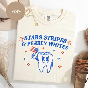 May include: Ivory-colored t-shirt with the text "STARS STRIPES & PEARLY WHITES" in blue. A cartoon tooth holding an American flag is featured in the design. The shirt is part of a patriotic collection.