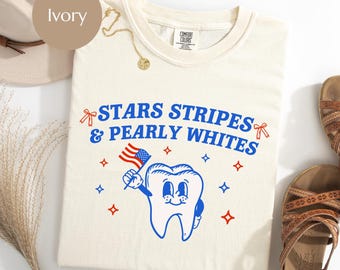 May include: Ivory-colored t-shirt with the text "STARS STRIPES & PEARLY WHITES" in blue. A cartoon tooth holding an American flag is featured in the design. The shirt is part of a patriotic collection.