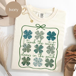 Peut inclure: T-shirt ivoire avec une bordure verte ornée de neuf trèfles à quatre feuilles dans des tons de bleu et de vert. Le texte "I teach the cutest clovers in the patch" est écrit en police script. Un collier doré est visible.
