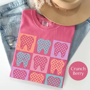 May include: A pink t-shirt with a grid pattern of colorful teeth. Each tooth is a different color, including orange, blue, and purple, with a checkered design. The shirt is folded on top of blue jeans, with a hat and necklace visible.