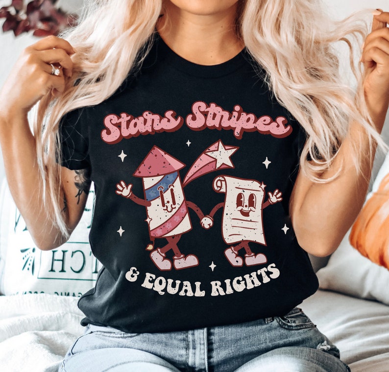 May include: Black t-shirt featuring a retro design with the text "Stars Stripes & Equal Rights." The graphic includes cartoon illustrations of a firework and a scroll holding hands, with red, white, and blue accents.