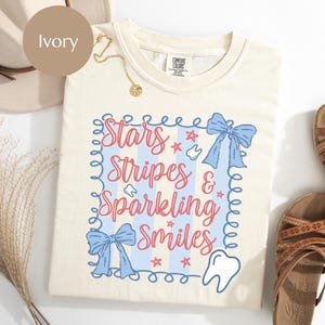 May include: Ivory-colored t-shirt with the text "Stars, Stripes & Sparkling Smiles" in red and blue. The shirt features a blue bow and star graphics. A gold necklace and sandals are also visible.