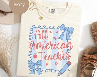 May include: Ivory-colored t-shirt with the text "All American Teacher" in red script, surrounded by blue bows, stars, and a pencil. The shirt has a crew neck and is made of Comfort Colors material.