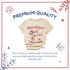 May include: Cream-colored t-shirt with rolled sleeves. The shirt features a graphic of a cartoon firework and a document holding hands, with the text "Stars Stripes & Equal Rights". The words "Premium Quality" are above the graphic.