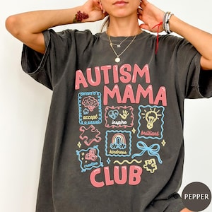 Op de afbeelding: Donkergrijs t-shirt met de tekst "AUTISM MAMA CLUB" in roze. Het shirt heeft verschillende illustraties, waaronder een hersen, hart, puzzelstukjes en een regenboog, met woorden als "accepteren", "inspireren" en "briljant".