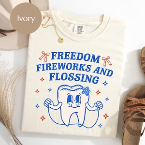 May include: Ivory-colored t-shirt with the text "FREEDOM FIREWORKS AND FLOSSING" in blue. A cartoon tooth character with a blue bow is featured below the text. Red bows and stars accent the design. The shirt is paired with sandals and a hat.