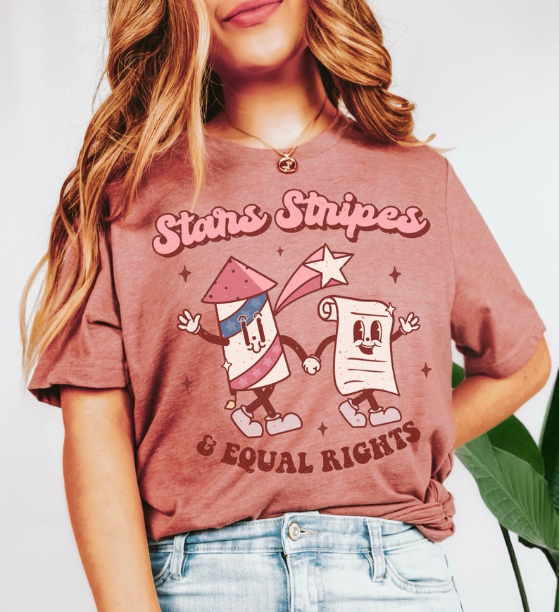 May include: A mauve-colored t-shirt with the text "Stars Stripes & Equal Rights" in a retro font. The graphic features a cartoon firework and a document holding hands, with a shooting star and small stars.