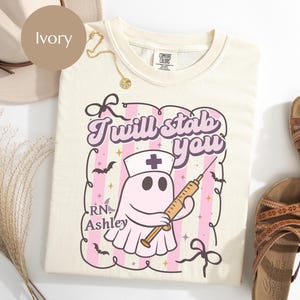 May include: An ivory t-shirt featuring a graphic of a ghost wearing a nurse cap and holding a syringe. The text reads "I will stab you" and "RN Ashley" over a pink and white striped background with small bats and stars inside a decorative frame.