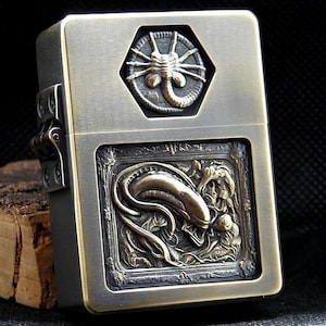 Könnte beinhalten: Ein messingfarbenes Feuerzeug mit detaillierten Reliefschnitzereien. Die Oberseite zeigt ein sechseckiges Design mit einem Alien-Gesicht, während die Frontplatte ein detailliertes Alien-Kreaturen-Design zeigt. Das Feuerzeug ist ein Sammlerstück.