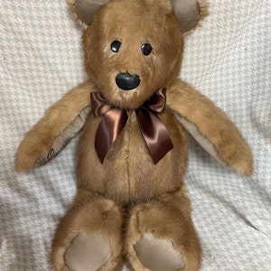 May include: A brown fur teddy bear with a brown satin bow. The bear has black eyes and a black nose.