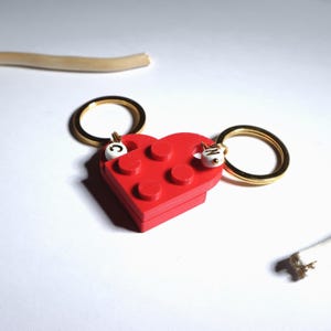 Pode incluir: Um porta-chaves vermelho em forma de coração feito de blocos interligados, com argolas douradas. O porta-chaves tem detalhes brancos com as letras "C" e "N".