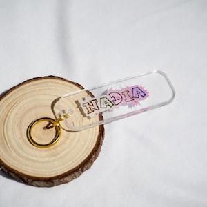 May include: A clear acrylic keychain with the name "NADIA" in a colorful, outlined font. The keychain is attached to a gold-colored ring. The keychain rests on a wooden slice with a light-colored background.