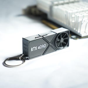 Può includere: Un portachiavi grigio con scheda grafica RTX 4090 con una ventola e la scritta "RTX 4090" in bianco. Il portachiavi è attaccato a un anello argentato. Un componente di computer argentato è sullo sfondo.