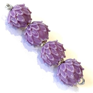 May include: A string of four lavender-colored glass beads, each shaped like a stylized flower bud. The beads are connected with silver-colored metal findings. The beads have a glossy finish and are set against a white background.