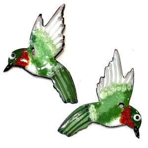 Könnte beinhalten: Zwei handgefertigte Kolibri-Ornamente. Jeder Vogel hat einen grünen Körper mit weißen und roten Akzenten und detaillierten weißen Flügeln. Die Vögel sind im Flug dargestellt, mit glänzendem Finish. Ideal für Naturliebhaber.