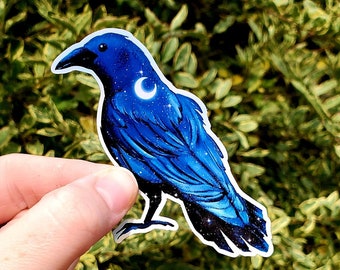 May include: A blue crow sticker with a crescent moon and stars on its feathers. The sticker is on a green leafy background.