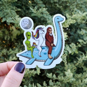 May include: A sticker featuring a blue dinosaur with a green alien, a unicorn, and a bigfoot riding on its back. The dinosaur is facing right and has a long neck. The unicorn is white with a rainbow mane and tail. The alien is green with large eyes. The bigfoot is brown and hairy. The sticker is on a white background.