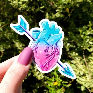 May include: A sticker of a pink and blue heart with a glitter effect, pierced by an arrow. The arrow is blue and purple with a white outline.