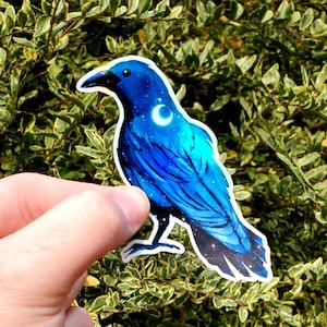 May include: A blue crow sticker with a crescent moon on its chest. The crow is depicted with a starry night sky pattern on its feathers.