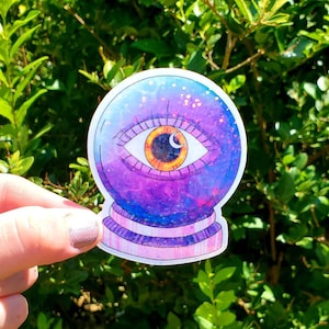 May include: A purple and blue glitter-filled glass orb with a large, golden eye inside. The eye has a crescent moon shape in the pupil.