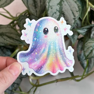 May include: A colorful sticker of a cartoon ghost with a rainbow gradient design. The ghost has a cute expression with large eyes and is surrounded by sparkling stars. The sticker is held in front of a green leafy plant.
