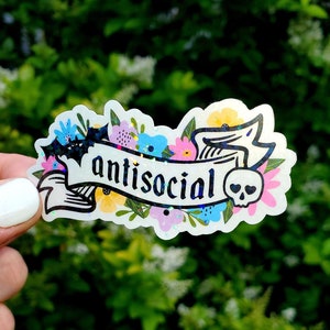May include: A clear sticker with a black banner that says "antisocial" with a skull and crossbones on the right side. The banner is surrounded by colorful flowers.