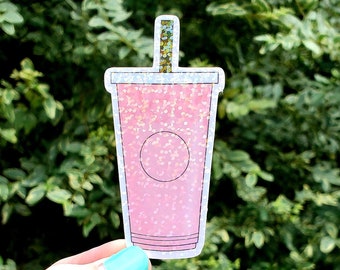 May include: Pink glitter sticker of a pink iced coffee cup with a straw. The sticker is on a hand.