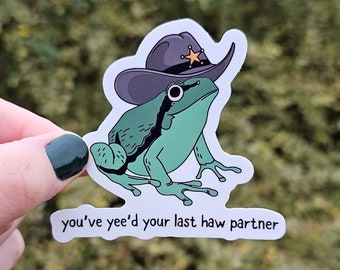 May include: A green frog wearing a sheriff's hat with a star on it. The frog is sitting on a white sticker with the text "you've yee'd your last haw partner".