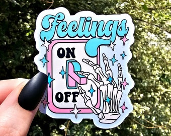 Può includere: Un adesivo con una mano scheletrica che aziona un interruttore etichettato "Feelings" con le opzioni "On" e "Off". L'interruttore è rosa e blu con stelle bianche.