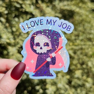 Op de afbeelding: Een holografische sticker met een cartoon Grim Reaper met een roze halo en de tekst "Ik hou van mijn werk".