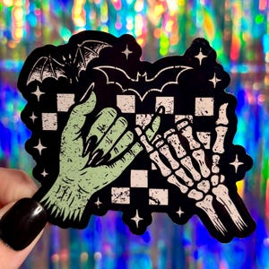 May include: A black sticker with a spooky theme. It features a green hand with long black nails holding hands with a skeleton hand. Bats and stars are above a checkered pattern.
