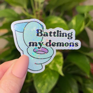 May include: A blue and white glitter sticker with a cartoon toilet and the text "Battling my demons".