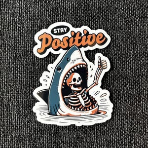 May include: A sticker featuring a cartoon shark with a skeleton inside, giving a thumbs-up. The shark is blue and white, with an open mouth. The words "Stay Positive" are above the shark in orange and white.