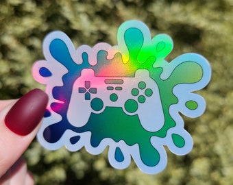 May include: A holographic sticker with a white outline of a video game controller in the center. The controller is surrounded by a colorful, iridescent splatter design.