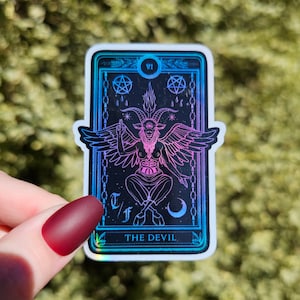 May include: A holographic tarot card sticker featuring the Devil card. The card is black with a pink and blue iridescent finish. The Devil is depicted as a goat-like figure with wings and a crown. The card also features the Roman numeral VI and the text "THE DEVIL".