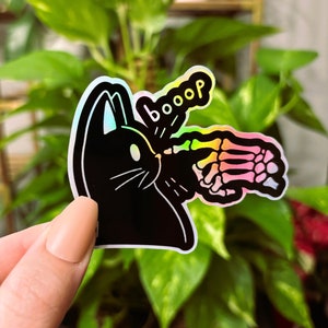 May include: A holographic sticker featuring a black cat with a skeleton hand reaching out and the word "booop" above it.
