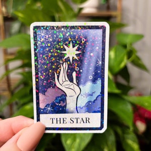 May include: A tarot card with a glittery blue background and a white hand reaching up to a white star. The card reads "THE STAR" in white text.
