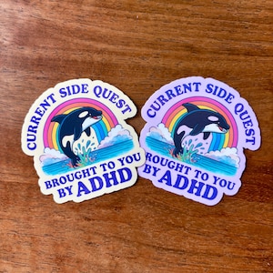 May include: Two stickers with a cartoon orca leaping from water, set against a rainbow. Text reads "Current Side Quest" and "Brought to you by ADHD" in a playful font. One sticker has a yellow background, the other is lavender.