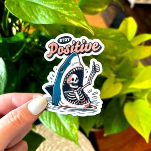 May include: A sticker featuring a cartoon shark with an open mouth, revealing a skeleton giving a thumbs-up. The sticker has the words "Stay Positive" in a stylized font, with a light pink and white color scheme. The shark is blue and white.