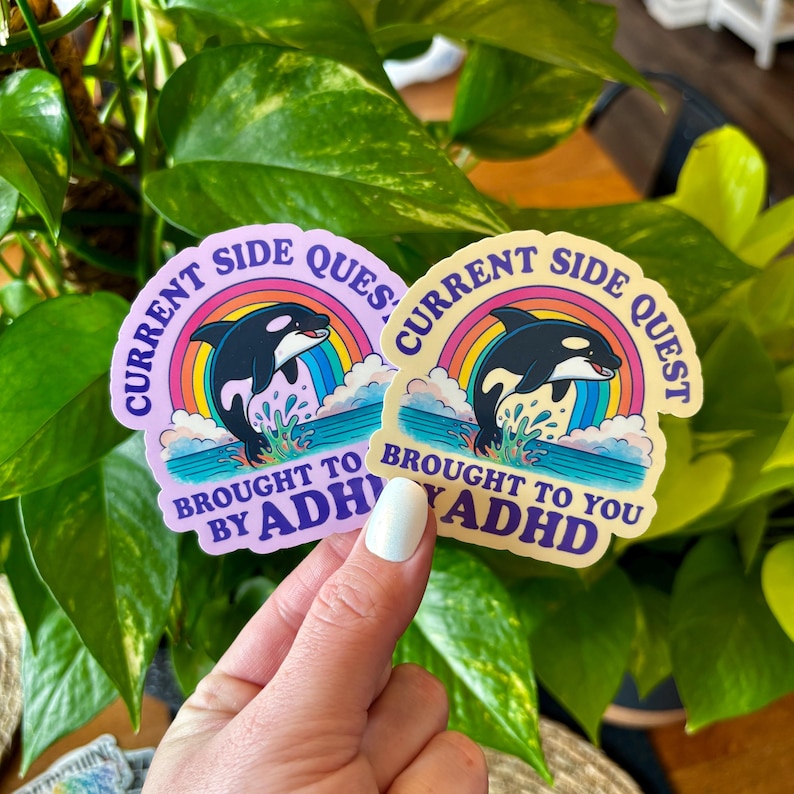 May include: Two stickers with a cartoon orca leaping from water under a rainbow. The stickers read "CURRENT SIDE QUEST" and "BROUGHT TO YOU BY ADHD". One sticker is light purple, the other is yellow.
