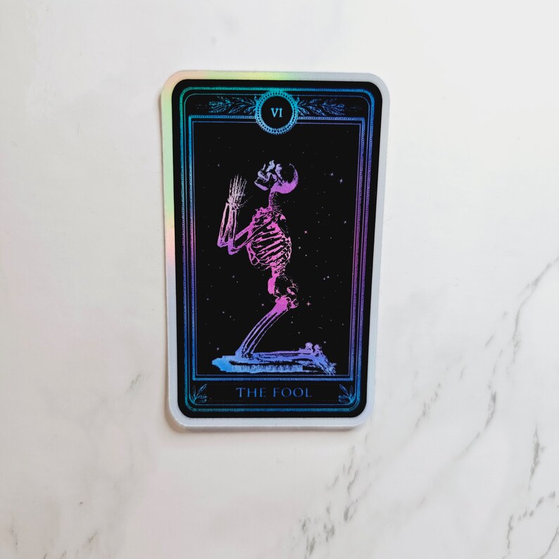 May include: A holographic tarot card with a black background and a purple and pink skeleton figure kneeling with its hands raised. The card is labeled "The Fool" and has the Roman numeral "VI" in a circle at the top.