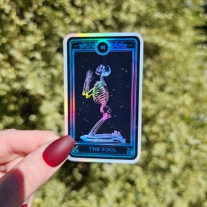 May include: A holographic tarot card with a black background and a rainbow skeleton figure kneeling with hands raised. The card is labeled "The Fool" and has the Roman numeral "VI" in the top right corner.